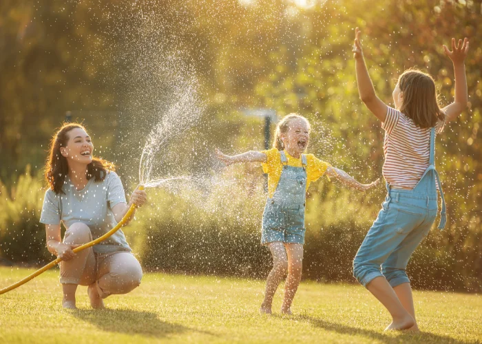 A mother spraying a garden hose while her daughters laugh and dance in the water on a sunny lawn.