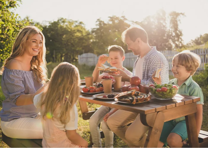 A smiling family enjoying a sunny outdoor picnic at a wooden table in a green backyard setting.