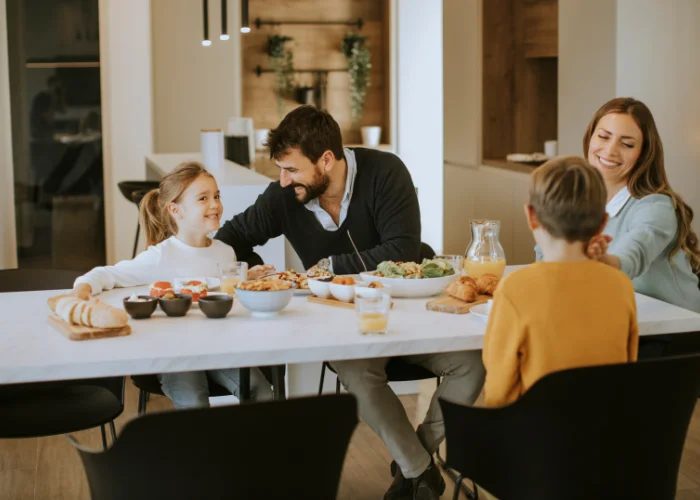 A happy family of four sitting around a modern dining table sharing a healthy breakfast together inside.