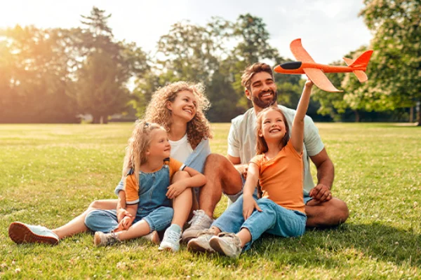 Happy parents sitting on a lush park lawn with their young daughters playing with a toy airplane.