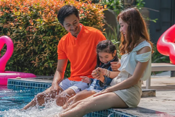 A family sitting poolside together with their feet in the water, smiling and splashing on a sunny day.