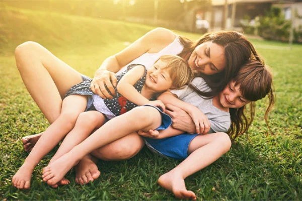 A smiling mother holding her two young children while relaxing together on a sunny green lawn.