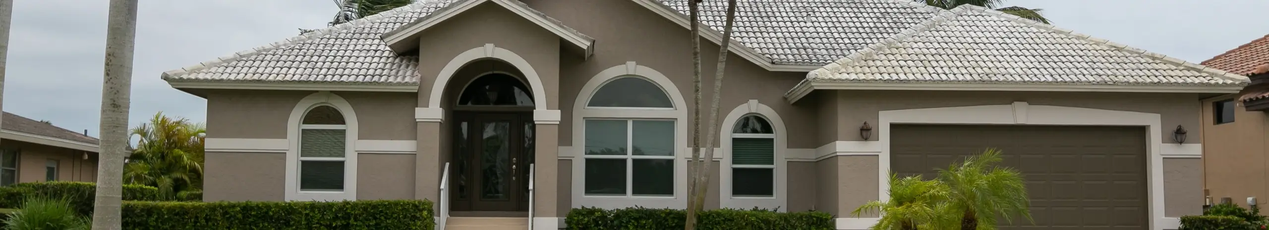 Exterior of a modern single-story taupe stucco home featuring a white tile roof, arched windows, and a brown garage door.