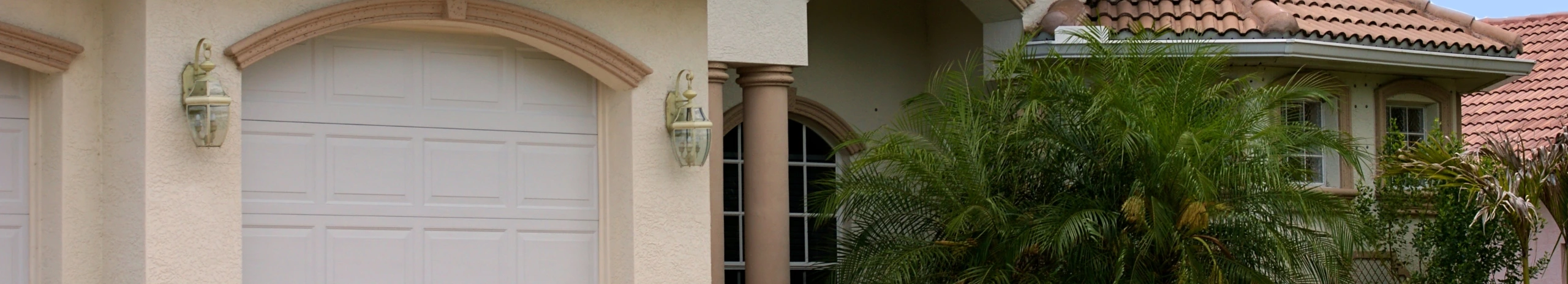 White paneled garage door on a light stucco home with a terracotta tile roof and palm landscaping.