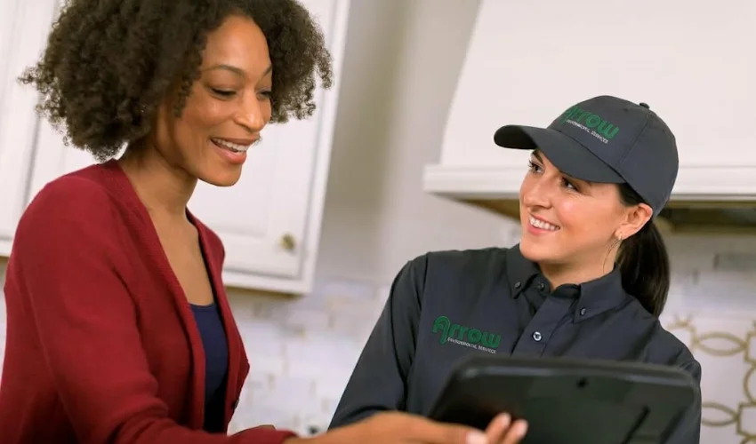 An Arrow Environmental pest control technician reviewing service details on a tablet with a smiling homeowner inside a kitchen.